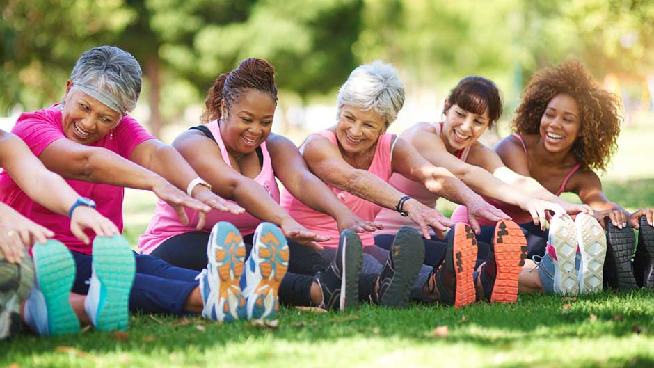 Diverse group of women wearing pink shirts and stretching in a row outdoors on a lawn