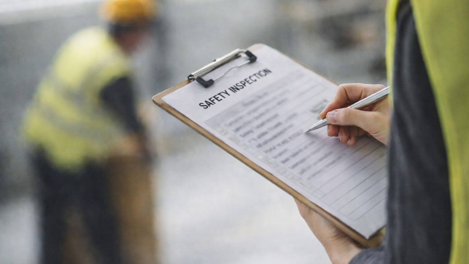 Blurred background and focus on a paper labeled Safety Inspection attached to a clipboard, clipboard held by a person wearing a yellow construction vest