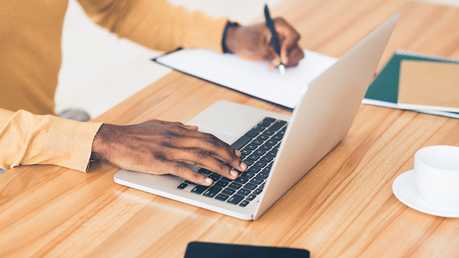 A person with one hand on a laptop and another writing with a pen